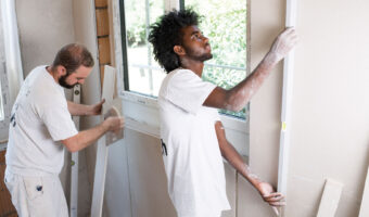 Ein Mann prüft mit einer Wasserwaage die vertikale Ausrichtung einer Wand, während ein zweiter Mann ein Holzstück vorbereitet.