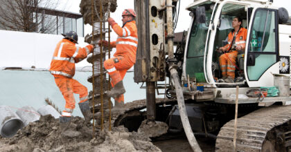 Auf der Baustelle verlassen zwei Männer einen Erdhaufen, während ein dritter Mann einen Erdbohrer bedient.