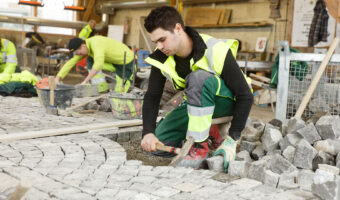 Ein Mann schlägt mit einem Pflästererhammer Pflastersteine in die Unterlage, während er auf einem niedrigen einbeinigen Stuhl sitzt.