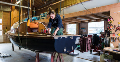 Eine Frau kniet auf einem kleinen hängenden Boot und befestigt im hinteren Teil des Bootes ein Seil.