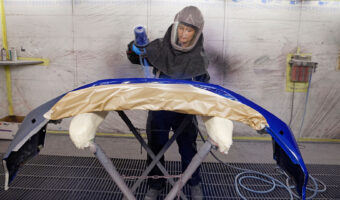 Eine Frau in einem Schutzanzug lackiert mit einer Spritzpistole die Stossstange eines Fahrzeugs.