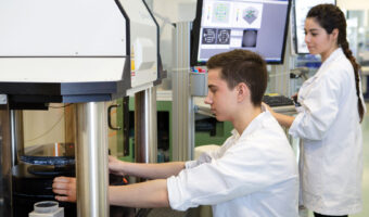 Ein Mann fertigt mit einer Fertigungsmaschine ein Glasprodukt, während eine Frau neben ihm einen Computer bedient.