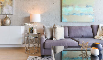 In einem Raum mit roher Betonwand stehen Sofa, Sofatisch und Beistelltische sowie Vasen und Lampen, an der Wand hängt ein abstraktes Bild.