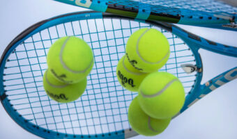 Drei neongelbe Tennisbälle liegen auf einem Spiegel in dem sich ein Tennisschläger spiegelt.