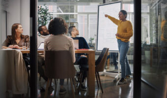 Eine Frau steht an einer Flipchart und zeigt darauf während zwei Frauen und zwei Männer an einem Tisch sitzend ihr dabei zu sehen.
