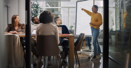 Eine Frau steht an einer Flipchart und zeigt darauf während zwei Frauen und zwei Männer an einem Tisch sitzend ihr dabei zu sehen.