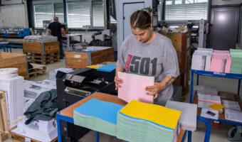 Eine Frau steht an einem Rollwagen und sortiert farbige Blätter zusammen.