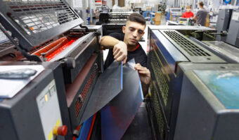 Ein Mann steht zwischen zwei Druckmaschinen und entfernt an der einen zwei Abdeckungen.