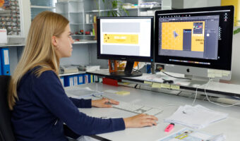 Eine Frau sitzt vor zwei Bildschirmen und layoutet eine Etikette für ein Honigglas.