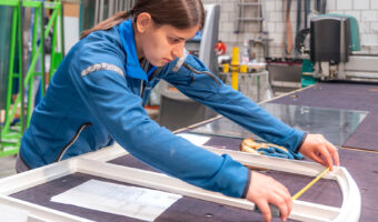 Eine Frau misst in einer Werkstatt den Fensterrahmen aus, der vor ihr auf dem Tisch liegt.