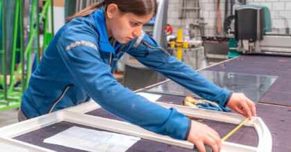 Eine Frau misst in einer Werkstatt den Fensterrahmen aus, der vor ihr auf dem Tisch liegt.