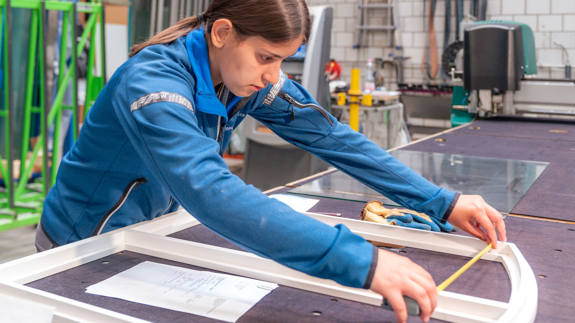 Eine Frau misst in einer Werkstatt den Fensterrahmen aus, der vor ihr auf dem Tisch liegt.