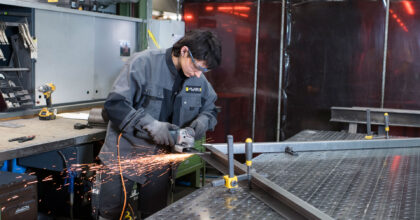 Eine Frau mit Schutzbrille steht an einem Tisch und flext an einem Metallrahmen.