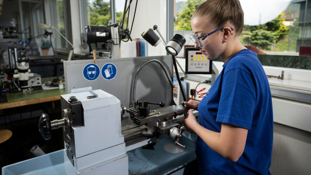Eine Frau mit Schutzbrille steht vor einer Maschine und stellt ein Metallteil her.
