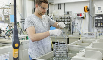 Ein Mann zieht ein Gestell mit kleinen Metallgegenständen aus einem chemischen Bad.