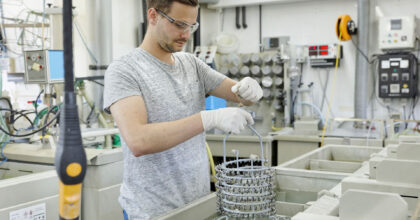 Ein Mann zieht ein Gestell mit kleinen Metallgegenständen aus einem chemischen Bad.