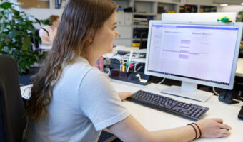 Eine Frau sitzt vor dem Computerbildschirm und führt mit der rechten Hand die Computermaus.