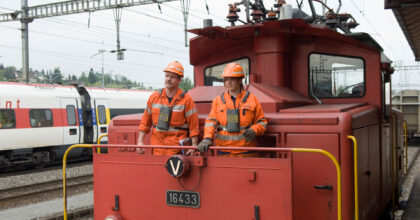 Eine Frau und ein Mann stehen auf einer Rangierlok und sehen auf die Geleise.