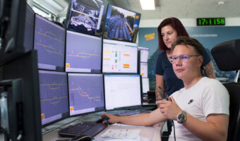 Ein Mann sitzt an einem Arbeitsplatz mit mehreren Bildschirmen, eine Frau steht hinter ihm.