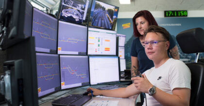 Ein Mann sitzt an einem Arbeitsplatz mit mehreren Bildschirmen, eine Frau steht hinter ihm.