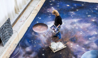 Eine Frau die in Socken auf einem grossen Bild am Boden steht, hält in der einen Hand die verkleinerte Vorlage des Bildes und in der anderen Hand einen Pinsel mit sehr langem Stiel und malt.