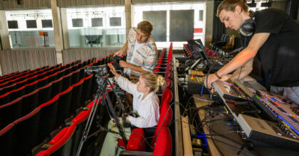 In einem Saal installieren eine Frau und ein Mann eine Kamera. Im Hintergrund richtet ein weiterer Mann ein Mischpult ein.