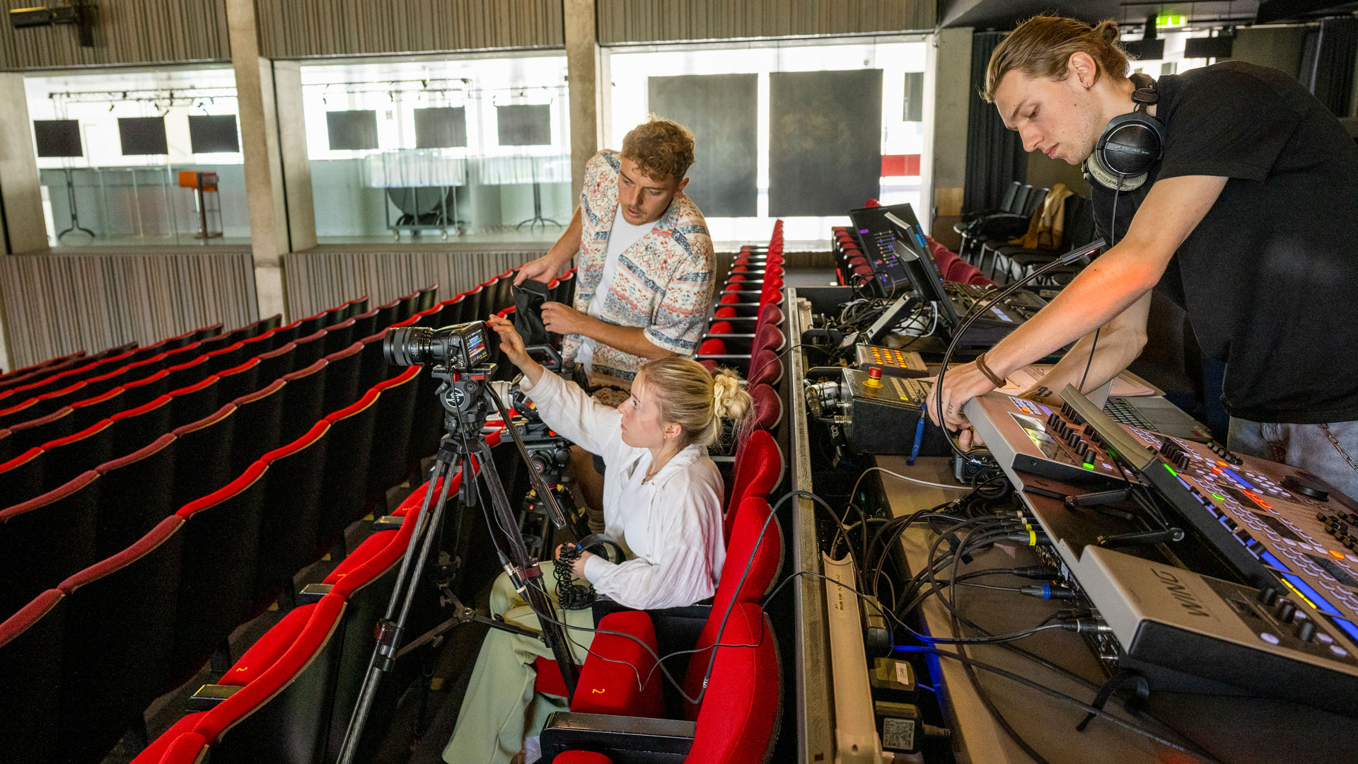 In einem Saal installieren eine Frau und ein Mann eine Kamera. Im Hintergrund richtet ein weiterer Mann ein Mischpult ein.