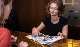 Eine Frau hinter einem Hoteldesk erklärt einem weiblichen Gast, anhand eines Planes, den Weg zu einer Sehenswürdigkeit.