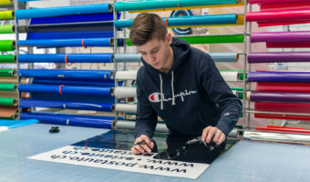 Ein junger Mann steht an einem Tisch, mit geplotteten Schriften, und zieht vorsichtig die überschüssige Folie ab.