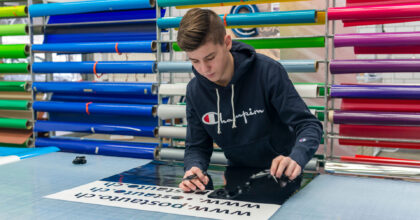 Ein junger Mann steht an einem Tisch, mit geplotteten Schriften, und zieht vorsichtig die überschüssige Folie ab.