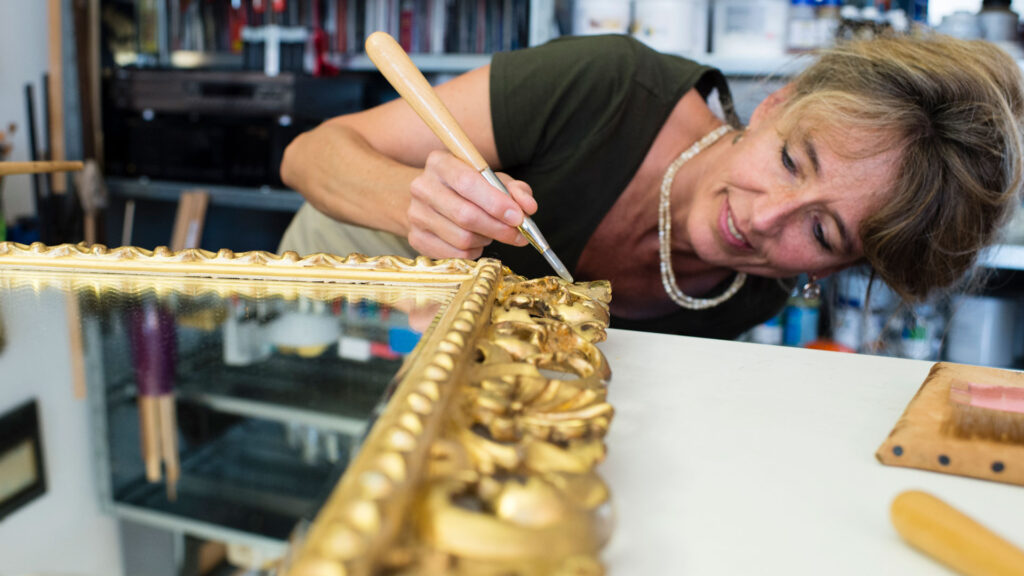 Eine Frau trägt mit einem Pinsel Goldfolie auf einen verschnörkelten Spiegelrahmen auf.