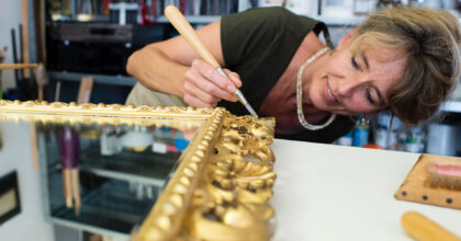Eine Frau trägt mit einem Pinsel Goldfolie auf einen verschnörkelten Spiegelrahmen auf.