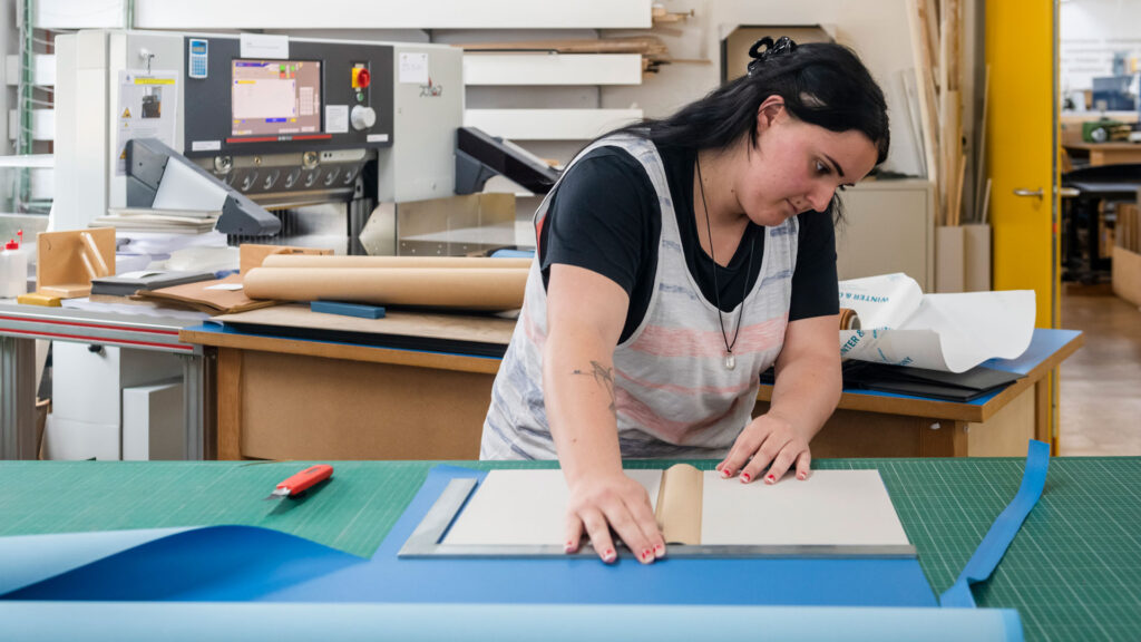 Eine Frau steht an einem Arbeitstisch und bereitet eine Buchhülle vor.