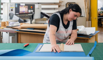 Eine Frau steht an einem Arbeitstisch und bereitet eine Buchhülle vor.