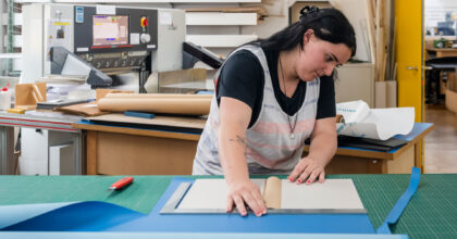 Eine Frau steht an einem Arbeitstisch und bereitet eine Buchhülle vor.