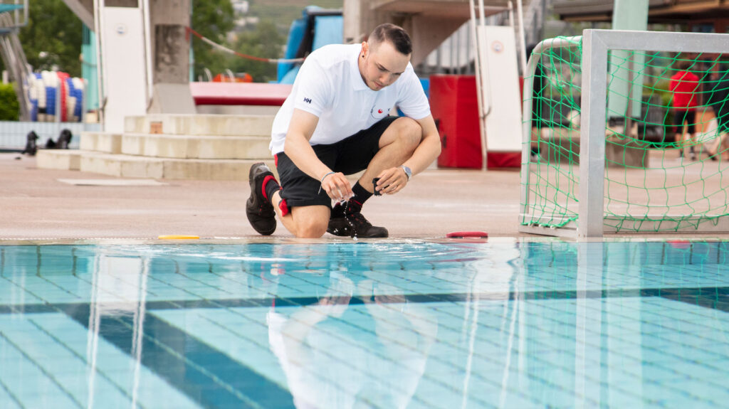 Ein Mann kniet an einem Schwimmbecken in einem Hallenbad und nimmt eine Wasserprobe.