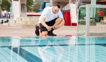 Ein Mann kniet an einem Schwimmbecken in einem Hallenbad und nimmt eine Wasserprobe.