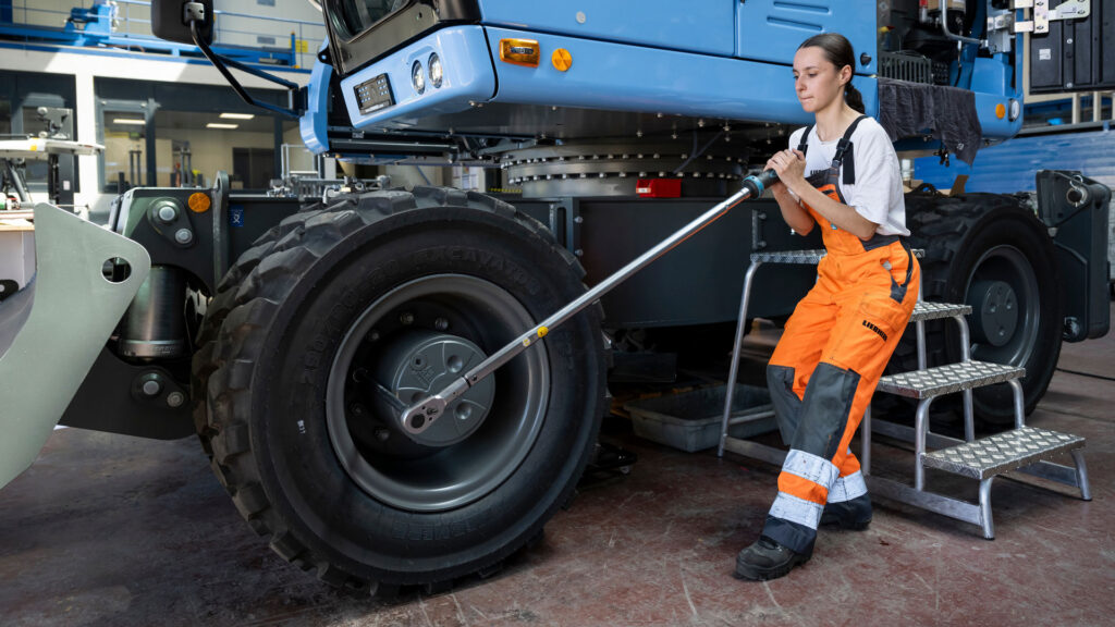 Eine Frau löst an einem Bagger die Radmutter mit einem langen Werkzeug.