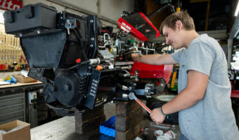 Ein lächelnder junger Mann hantiert am Motor eines Balkenmähers.