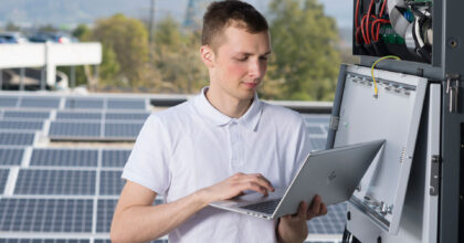 Ein junger Mann steht mit einem Laptop in den Händen auf einem Dach mit Solarpanels und prüft die Funktion.