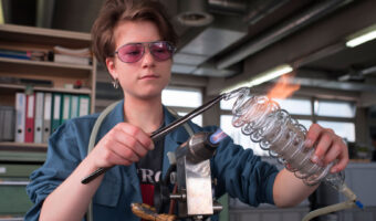 Eine junge Frau mit Schutzbrille erstellt eine Spirale aus Glas.