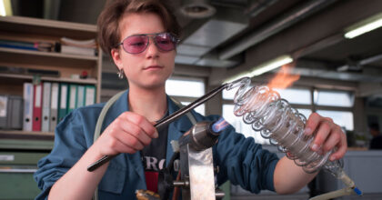 Eine junge Frau mit Schutzbrille erstellt eine Spirale aus Glas.
