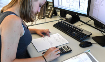 Eine junge Frau sitzt an einem Arbeitsplatz und rechnet mit einem Taschenrechner Masse aus.