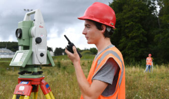 Ein junger Mann steht auf einer Wiese hinter seinem Messgerät und spricht in ein Funkgerät.