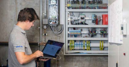 Ein Mann mit einem Laptop in den Händen steht vor einer geöffneten Kälteanlage und prüft die Funktion.